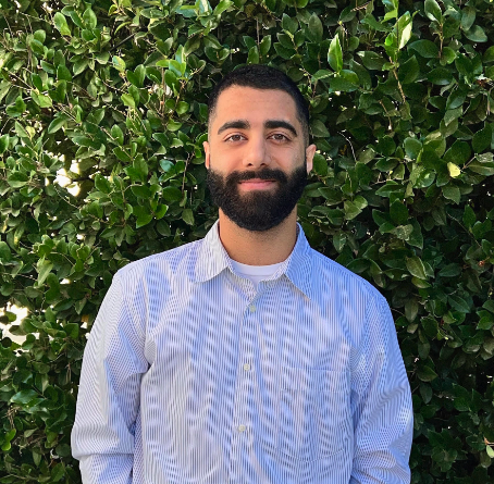 A man with a beard standing in front of bushes.